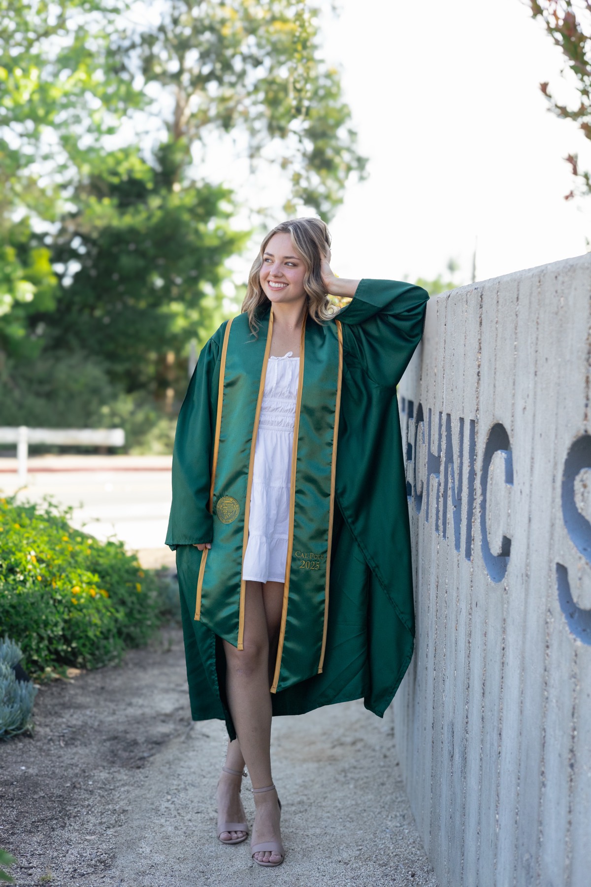 Graduate in a green gown leaning on the Polytechnic wall at Cal Poly