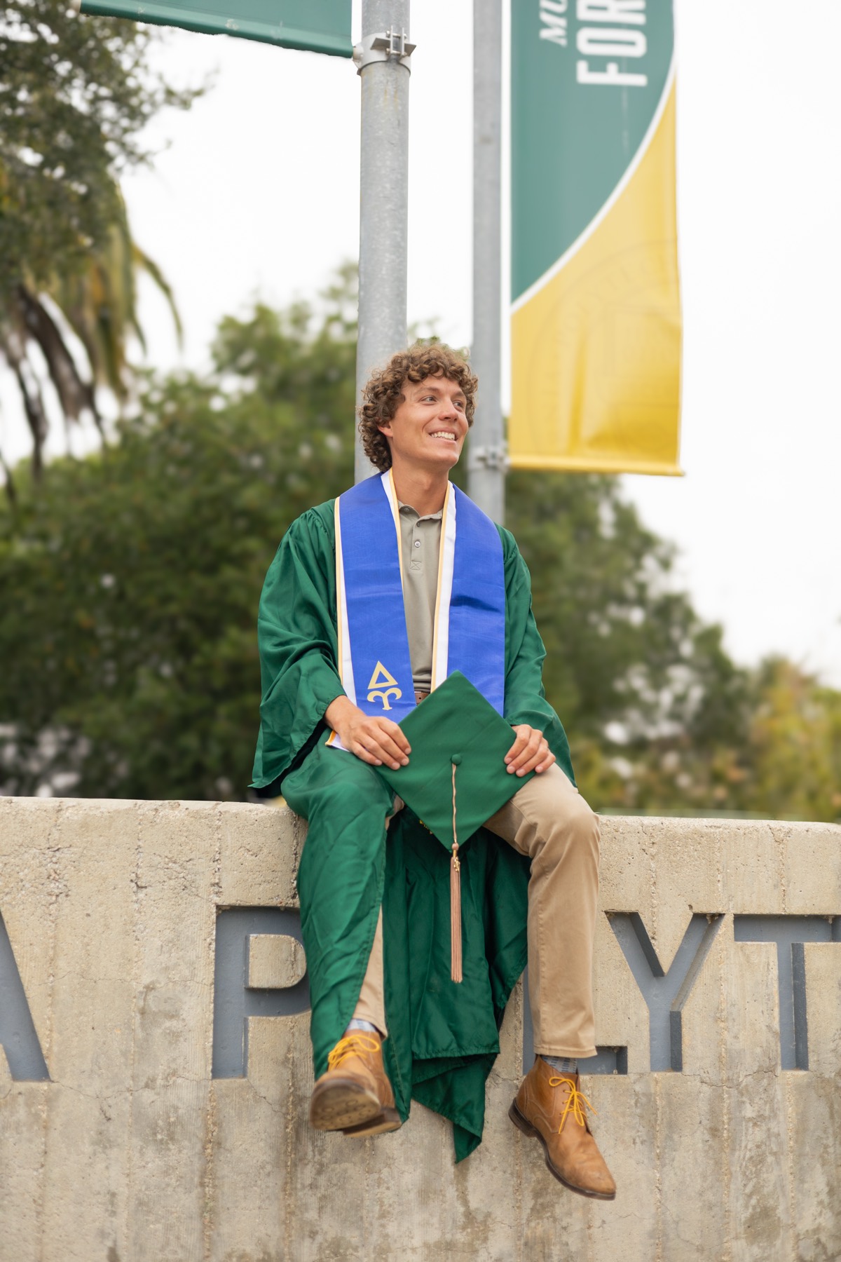 Graduate sitting on the Cal Poly sign holding his cap